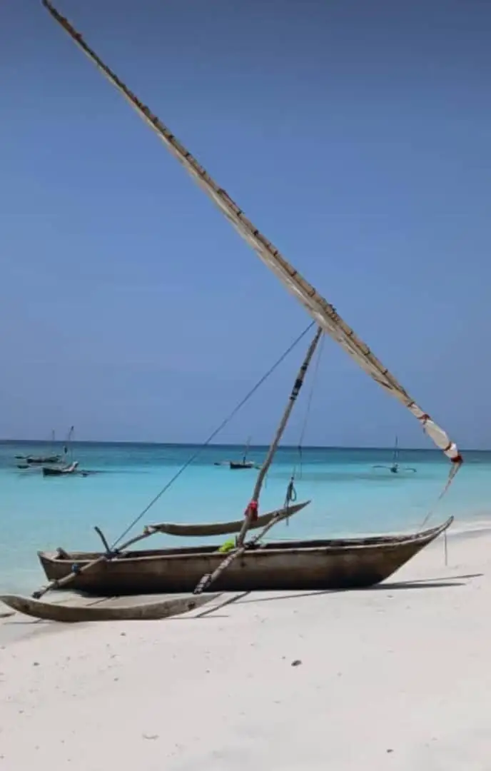 A traditional outrigger dhow drawn up on white sand with its sail spar raised, more boats moored beyond.