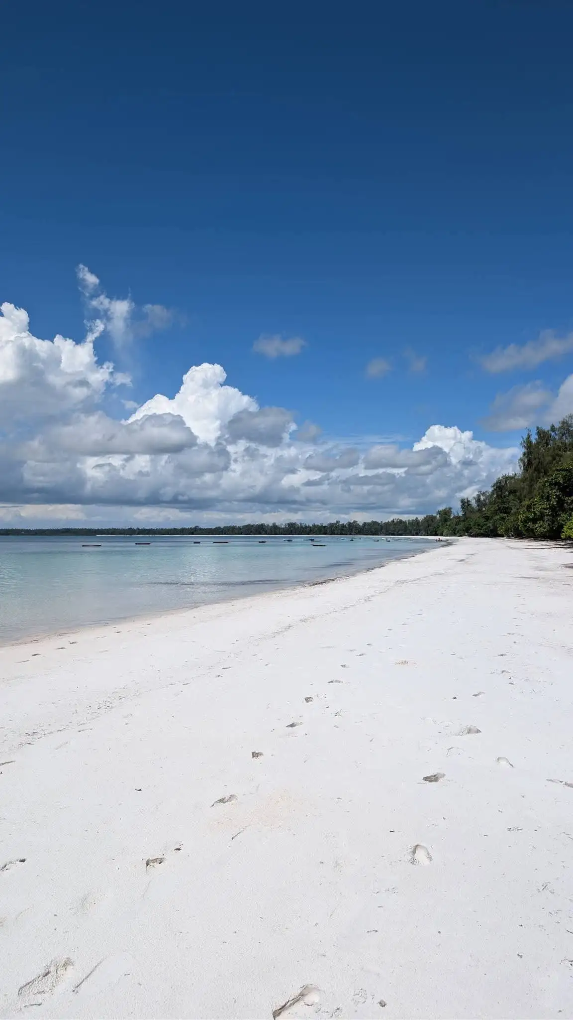 A long empty white beach under tall clouds, with boats out on the water.