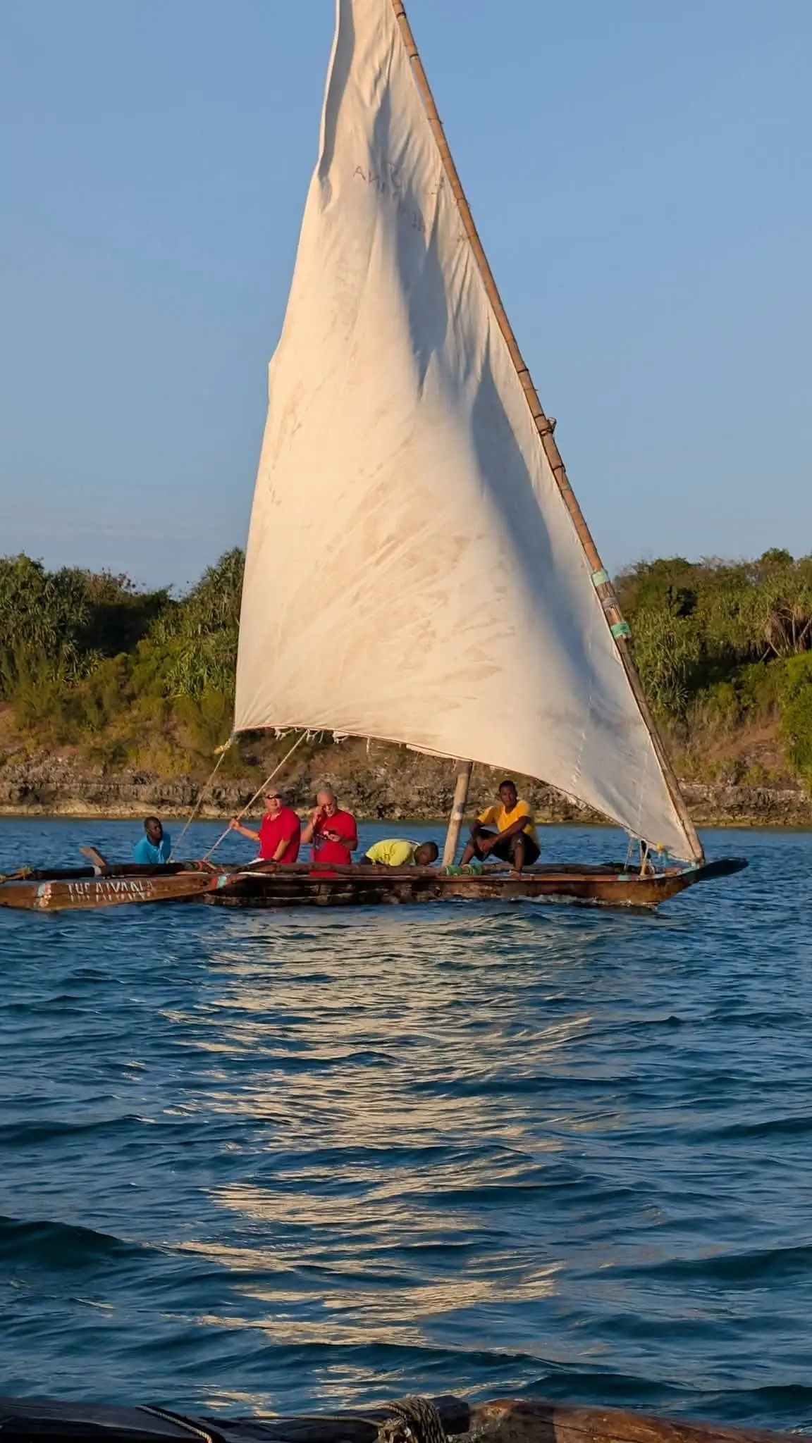 Guests sitting aboard a dhow as it sails under a tall white sail.