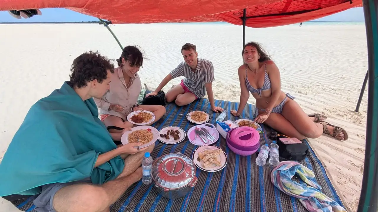 Four guests sitting on a mat under a red canopy, eating lunch on the sand.