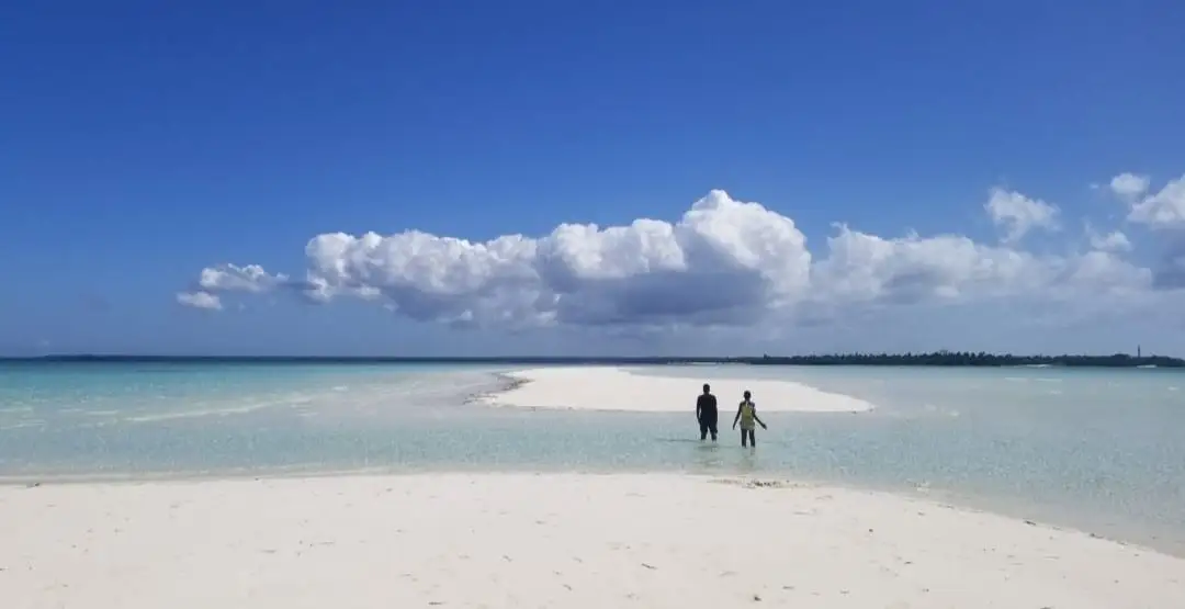A long white sandbank running out into turquoise water, with two people wading towards it.